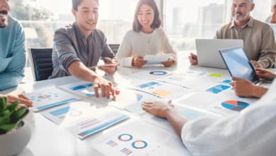 Sei persone discutono sorridenti intorno a un tavolo riguardo alcuni fogli con immagini di diagrammi